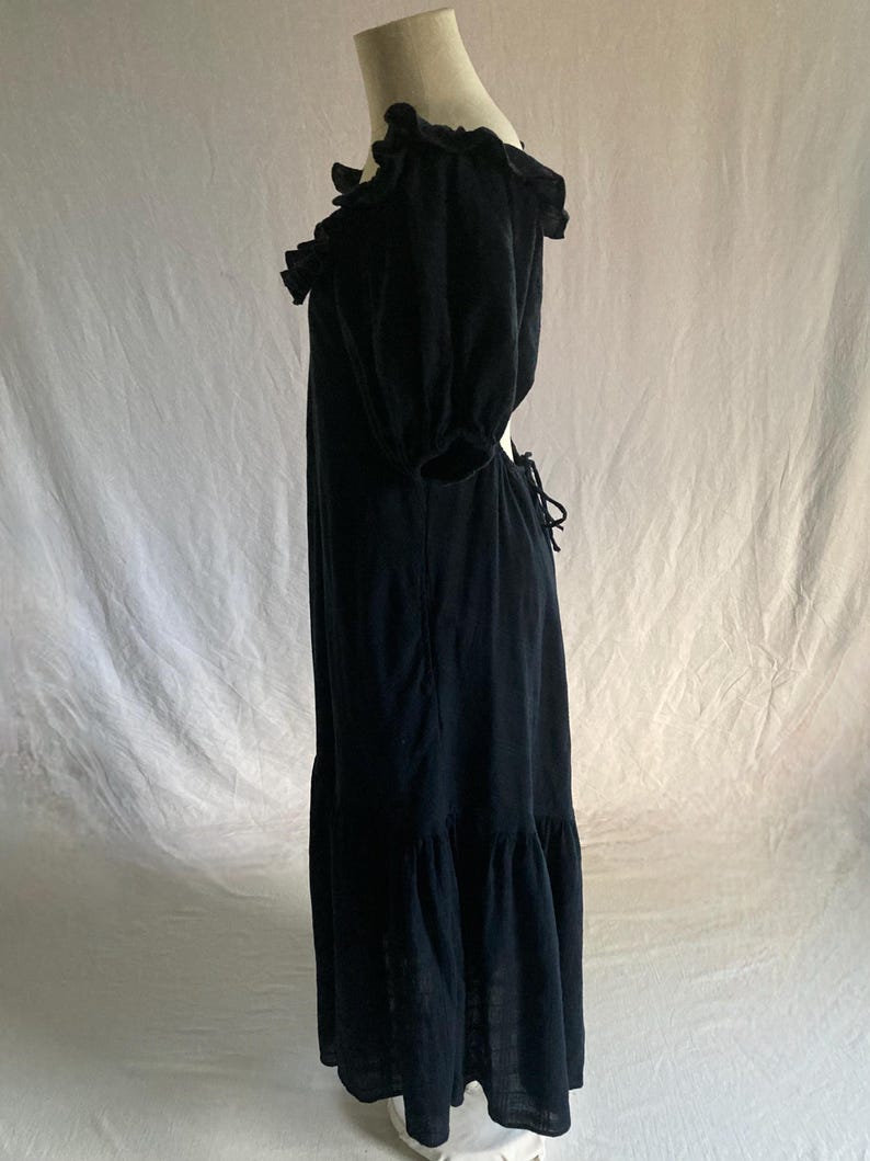 May include: A black maxi dress with a ruffled neckline and short, puffed sleeves. The dress features a tiered skirt and a tie-back detail. The fabric appears to be a lightweight material, suitable for warmer weather. The dress is displayed on a mannequin.