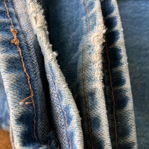 Pu&ograve; includere: Primo piano di jeans in denim blu chiaro con bordi usurati e cuciture arancioni visibili. Il tessuto mostra segni di usura, con dettagli sfilacciati lungo le cuciture, evidenziando un'estetica vintage o usurata. L'immagine si concentra sulla trama e sui dettagli del denim.