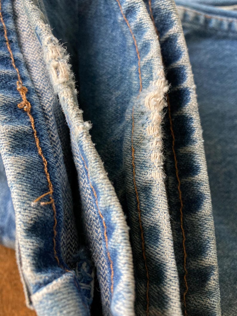 Pu&ograve; includere: Primo piano di jeans in denim blu chiaro con bordi usurati e cuciture arancioni visibili. Il tessuto mostra segni di usura, con dettagli sfilacciati lungo le cuciture, evidenziando un'estetica vintage o usurata. L'immagine si concentra sulla trama e sui dettagli del denim.