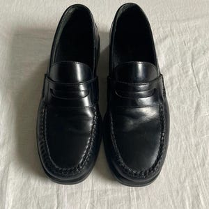 May include: A pair of black leather penny loafers. The shoes have a glossy finish, a stitched vamp, and a strap across the top. The soles are black and the shoes are displayed on a white surface. These classic shoes are suitable for various occasions.