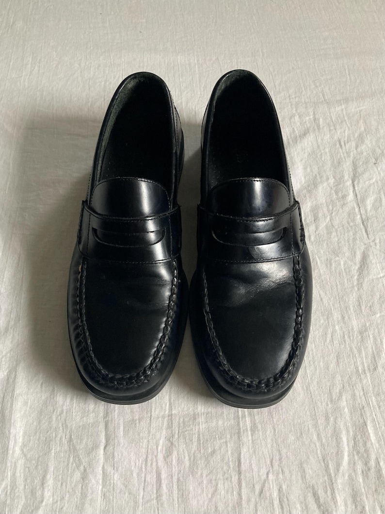 May include: A pair of black leather penny loafers. The shoes have a glossy finish, a stitched vamp, and a strap across the top. The soles are black and the shoes are displayed on a white surface. These classic shoes are suitable for various occasions.