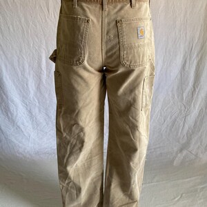 May include: Tan work pants with multiple pockets and a Carhartt logo patch on the back pocket. The pants have a straight leg design and a worn appearance. The pants are displayed on a mannequin.
