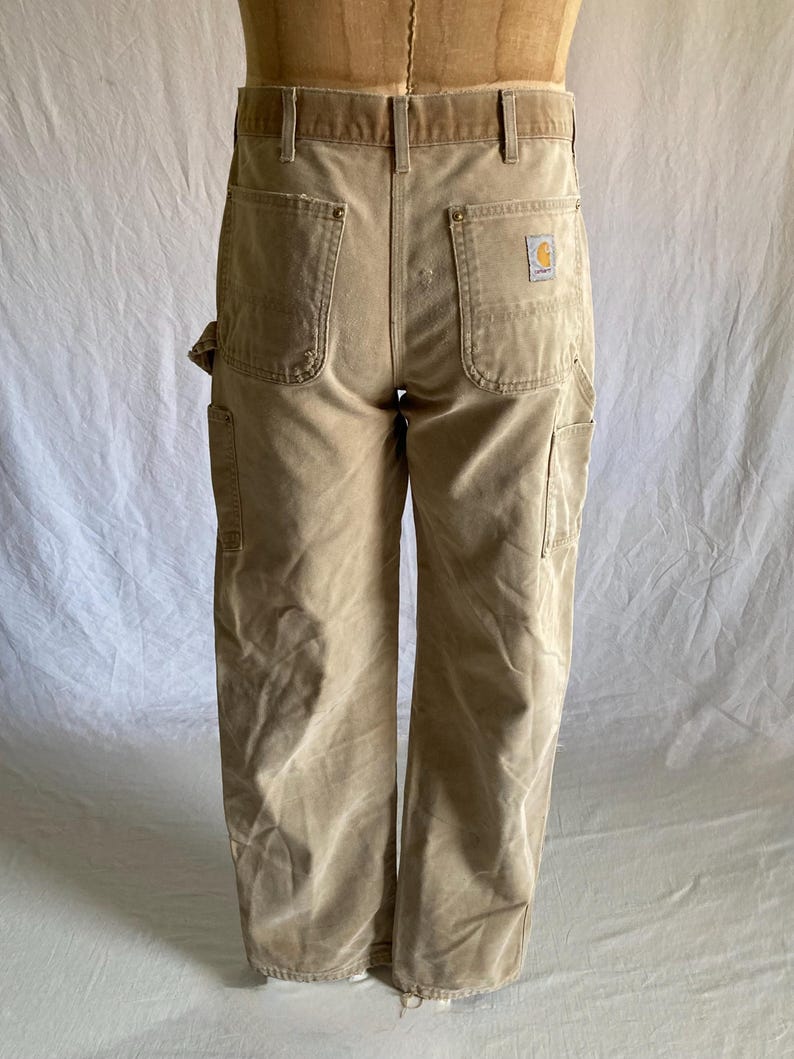 May include: Tan work pants with multiple pockets and a Carhartt logo patch on the back pocket. The pants have a straight leg design and a worn appearance. The pants are displayed on a mannequin.