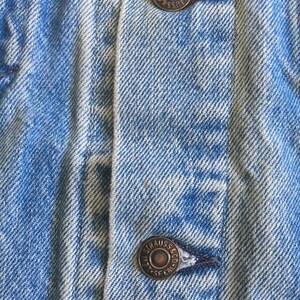 May include: Close-up of a blue denim jacket with two metal buttons. The buttons are round and have the text "Levi Strauss & Co. SF. Cal." engraved on them.