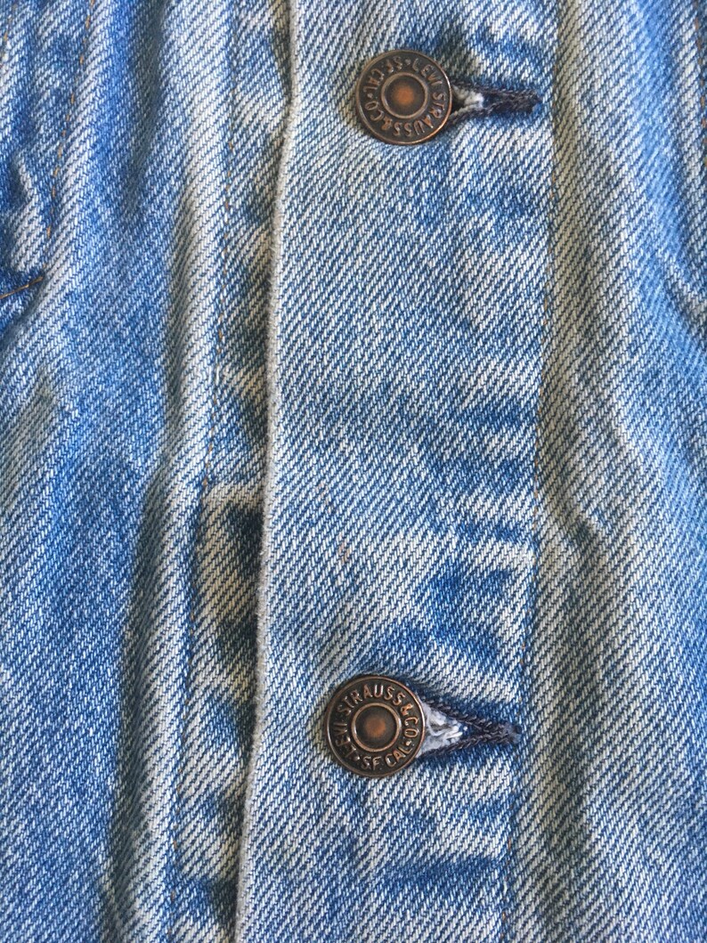 May include: Close-up of a blue denim jacket with two metal buttons. The buttons are round and have the text "Levi Strauss & Co. SF. Cal." engraved on them.