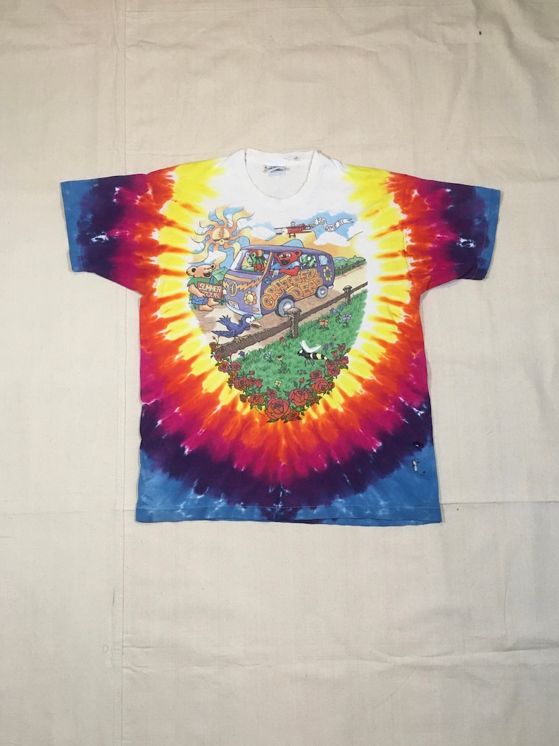 May include: A tie-dye t-shirt with a colorful swirl pattern. The shirt features a cartoon image of a van with the words "Grateful Dead" on the side. The van is surrounded by cartoon characters and flowers.