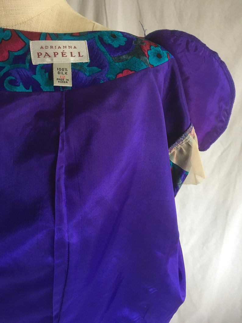 Op de afbeelding: Een paarse zijden jurk met een kraag met bloemenprint. Het label vermeldt "Adrianna Papell" en "100% Zijde" met maat 12 en "Gemaakt in Korea".