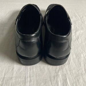 May include: A pair of black leather slip-on shoes. The shoes have a rounded toe and a low heel. The uppers are smooth and shiny, with a strap across the top of the foot. The shoes are sitting on a white surface.