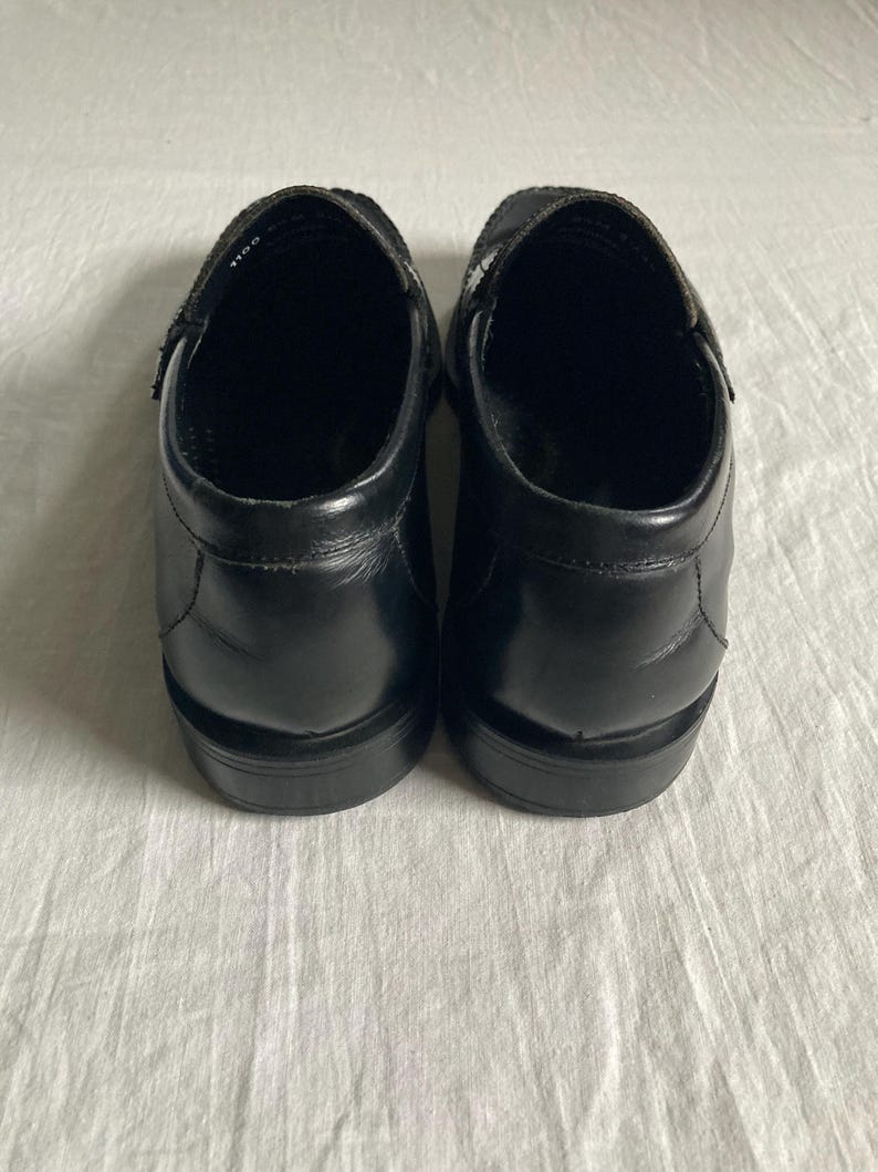 May include: A pair of black leather slip-on shoes. The shoes have a rounded toe and a low heel. The uppers are smooth and shiny, with a strap across the top of the foot. The shoes are sitting on a white surface.