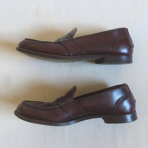 May include: A pair of brown leather loafers with a penny strap detail. The loafers have a slightly rounded toe and a low heel.