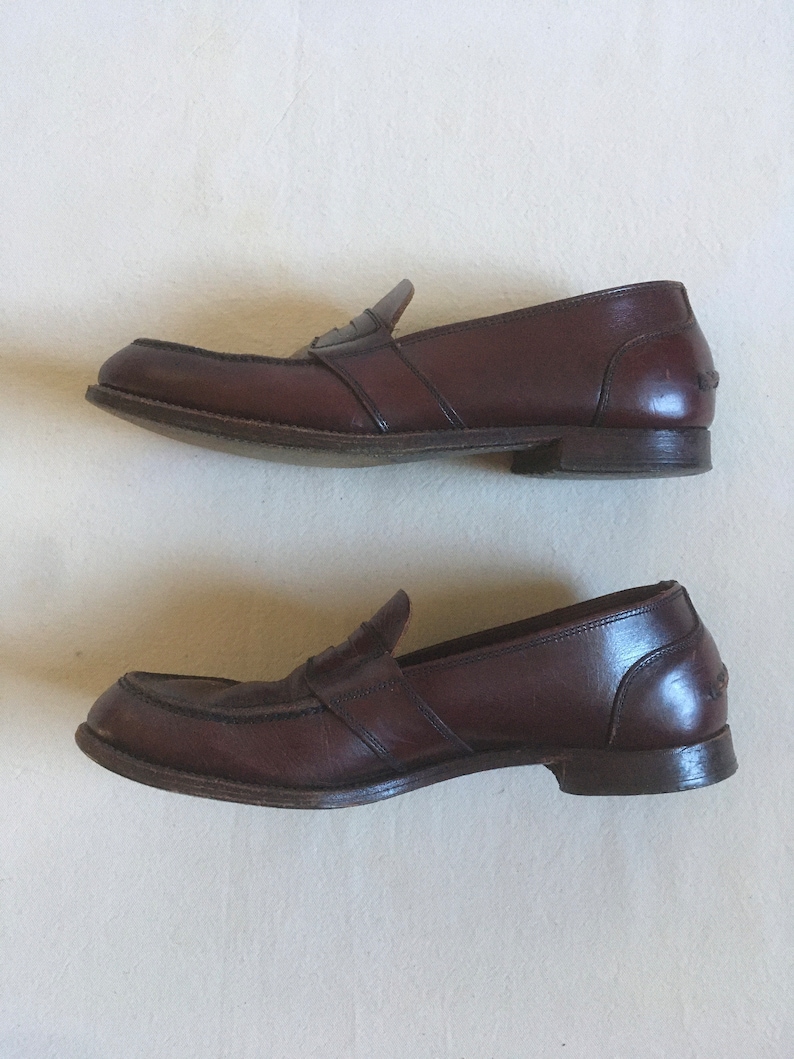 May include: A pair of brown leather loafers with a penny strap detail. The loafers have a slightly rounded toe and a low heel.