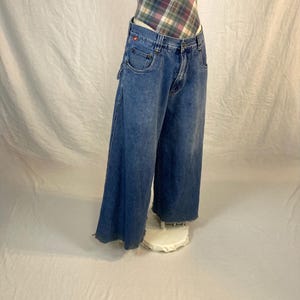 K&ouml;nnte beinhalten: Blaue Jeans mit weitem Bein aus Denim mit verwaschener Optik und ausgefranstem Saum. Die Jeans haben ein klassisches F&uuml;nf-Taschen-Design und ein rotes Logo-Detail auf der Vordertasche. Die Jeans werden auf einem wei&szlig;en St&auml;nder gezeigt.
