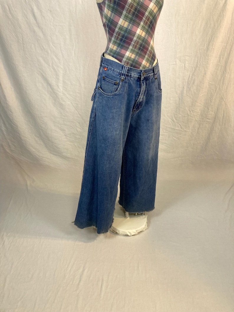 K&ouml;nnte beinhalten: Blaue Jeans mit weitem Bein aus Denim mit verwaschener Optik und ausgefranstem Saum. Die Jeans haben ein klassisches F&uuml;nf-Taschen-Design und ein rotes Logo-Detail auf der Vordertasche. Die Jeans werden auf einem wei&szlig;en St&auml;nder gezeigt.