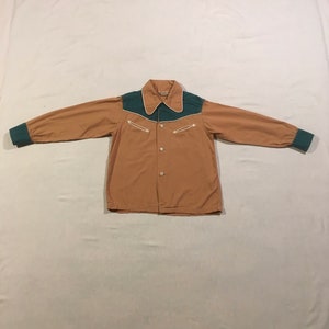 Puede incluir: Una camisa de manga larga de color marrón claro con un yugo verde y costuras blancas. La camisa tiene dos bolsillos en el pecho con costuras blancas y un cuello puntiagudo.