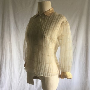 May include: A sheer, ivory-colored blouse with gold trim on the collar and cuffs. The blouse has a plaid pattern and a button-up front.