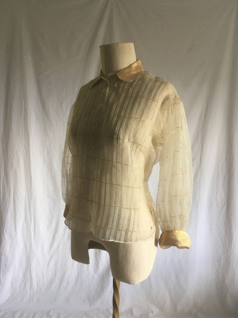 May include: A sheer, ivory-colored blouse with gold trim on the collar and cuffs. The blouse has a plaid pattern and a button-up front.