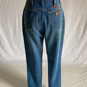 Pu&ograve; includere: Vista posteriore di jeans di denim blu a vita alta. I jeans hanno due tasche posteriori, una toppa in pelle marrone e un'etichetta rossa. I jeans sono esposti su un manichino.