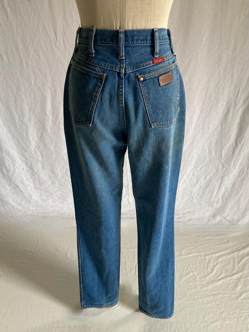 Pu&ograve; includere: Vista posteriore di jeans di denim blu a vita alta. I jeans hanno due tasche posteriori, una toppa in pelle marrone e un'etichetta rossa. I jeans sono esposti su un manichino.