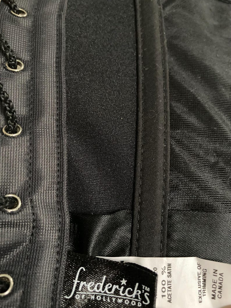 May include: Close-up of a black satin fabric label with white text that reads "frederick's of Hollywood" and "100% Acetate Satin". The label also includes the text "Exclusive of Trimming" and "Made in Canada".