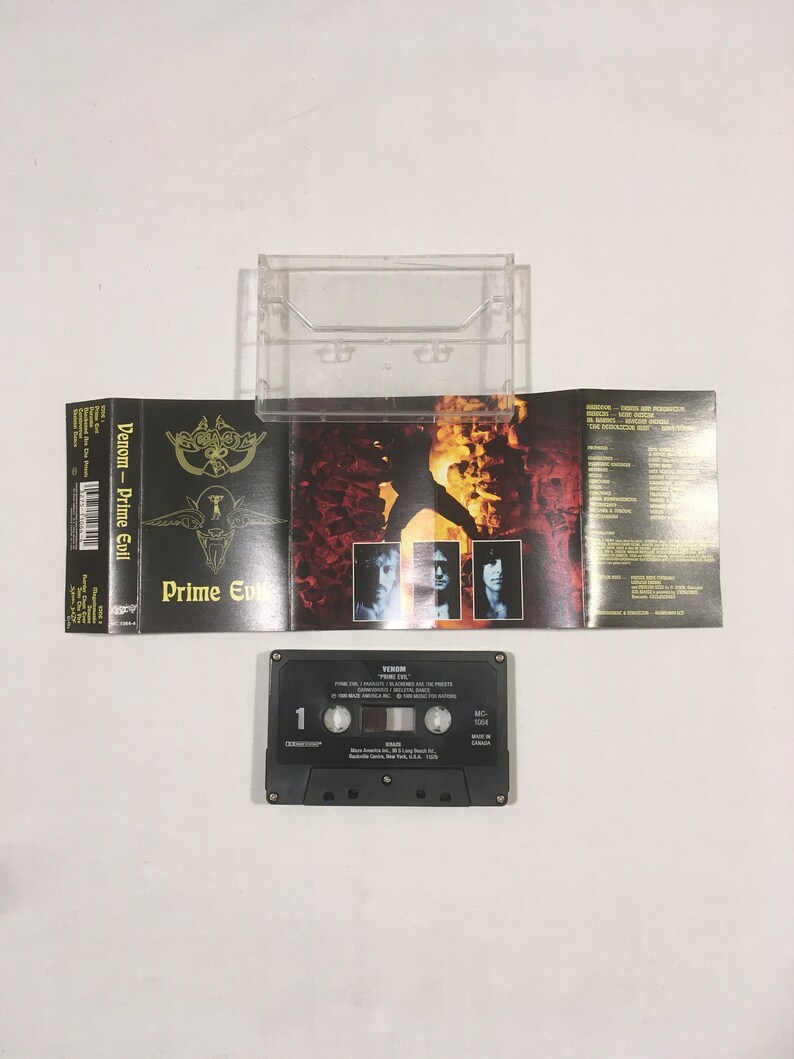 May include: A black cassette tape with the title "Venom - Prime Evil" on the label. The cassette tape is in a clear plastic case with a black and white album cover featuring a figure in a fiery background.
