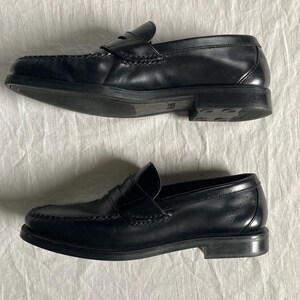 May include: A pair of black leather penny loafers. The shoes feature a classic design with a rounded toe, a strap across the vamp, and a low heel. The leather appears smooth and polished. The soles are dark.