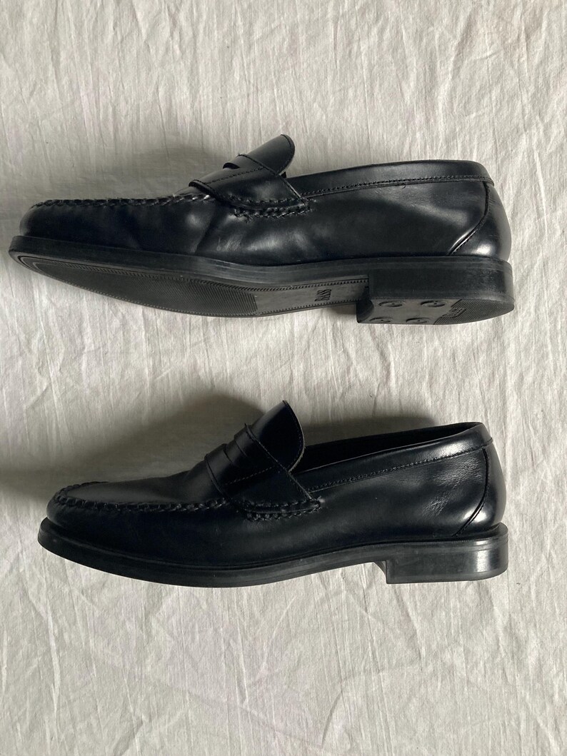 May include: A pair of black leather penny loafers. The shoes feature a classic design with a rounded toe, a strap across the vamp, and a low heel. The leather appears smooth and polished. The soles are dark.