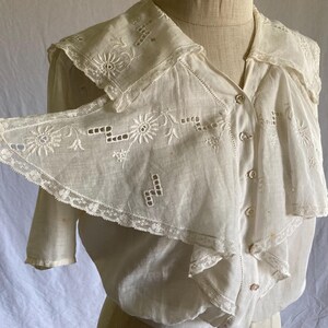 May include: A vintage white blouse with a large, detailed collar. The collar and front of the blouse feature intricate floral embroidery and delicate lace trim. The blouse has short sleeves and a button-down front, showcasing a classic, elegant design.
