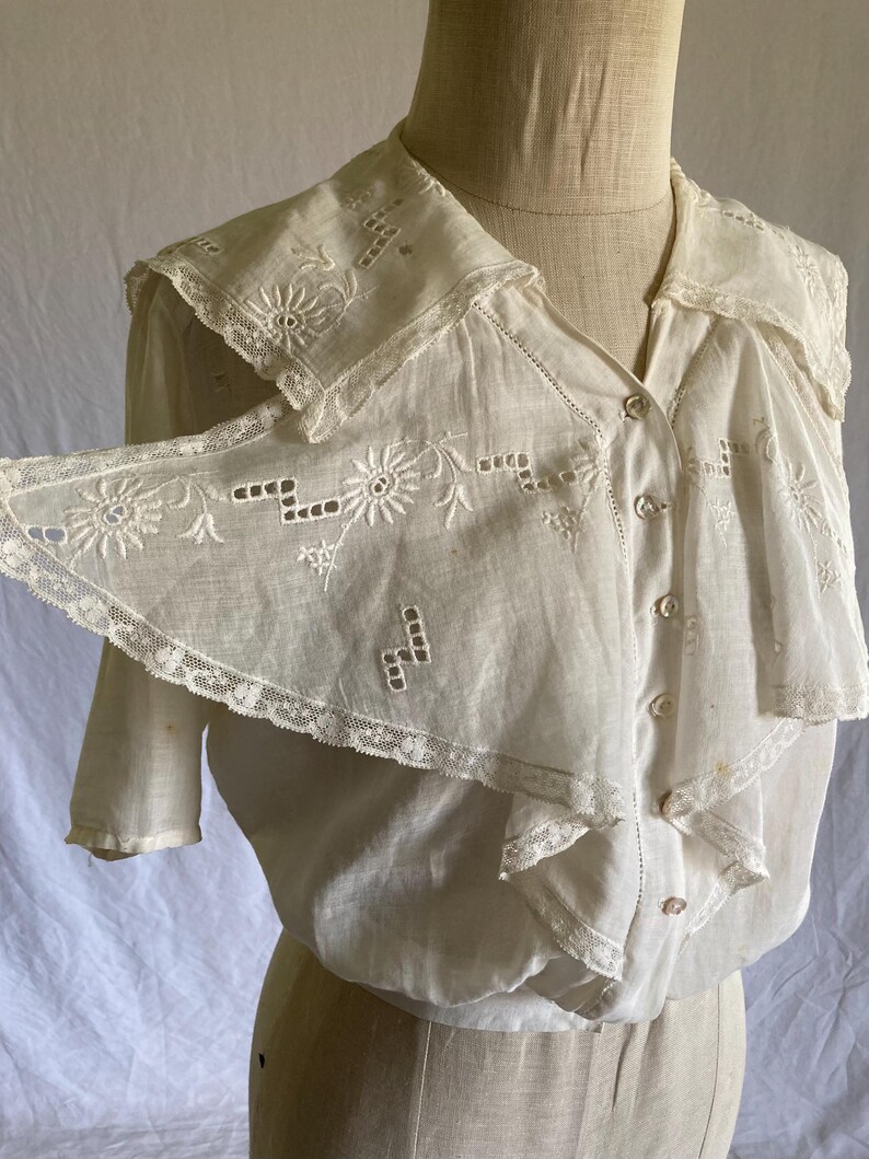 May include: A vintage white blouse with a large, detailed collar. The collar and front of the blouse feature intricate floral embroidery and delicate lace trim. The blouse has short sleeves and a button-down front, showcasing a classic, elegant design.