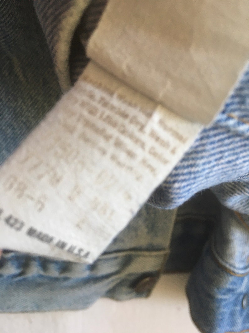 May include: Close-up of a light blue denim jacket with a white care label. The label has text that reads "Machine Wash Cold, Tumble Dry Low, Do Not Bleach, Do Not Iron, Do Not Dry Clean, Made in U.S.A."