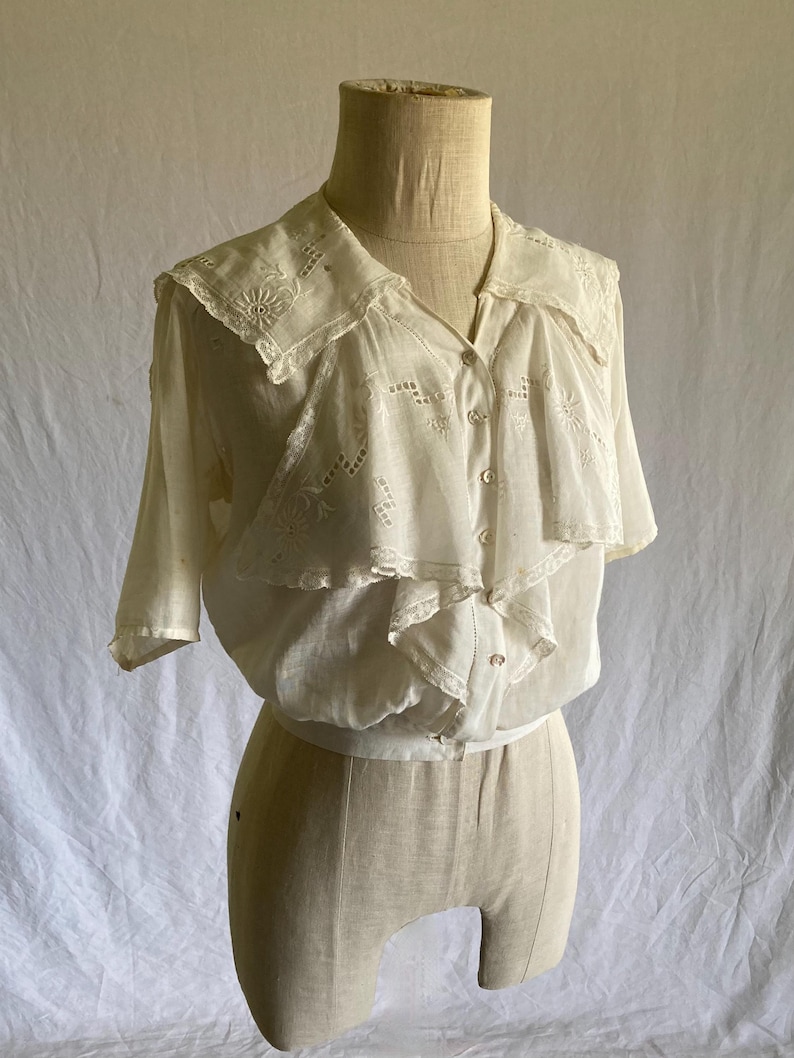May include: A vintage white blouse with short sleeves, a ruffled front, and a decorative collar. The blouse features delicate embroidery and button closures. The garment is displayed on a mannequin.