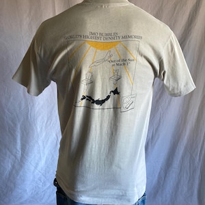 May include: A light gray t-shirt with a graphic of a plane flying through the sun. The text "IMO BUBBLES WORLD'S HIGHEST DENSITY MEMORIES" is printed above the graphic. The text "Out of the Sun at Mach 1" is printed below the graphic.