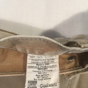 Puede incluir: Una etiqueta de ropa de trabajo Carhartt de color beige con texto blanco. La etiqueta incluye el nombre de la marca, la talla y las instrucciones de cuidado. El texto dice: "0804 08227 Carhartt Inc. P.O. Box 600 Dearborn MI 48126 USA Made in USA Fabriqu&eacute; aux E.U. Hecho en los EE.UU. 100% Cotton. Exclusive Trimming. 100% coton. Sane garniture. 100 algod&oacute;n. Sin incluir adorno.  Procter & Gamble  UREN Carhartt Cure on reverse side. Entretien au verso. Instrucciones de cuidado al dorso. B01 BRN 34"
