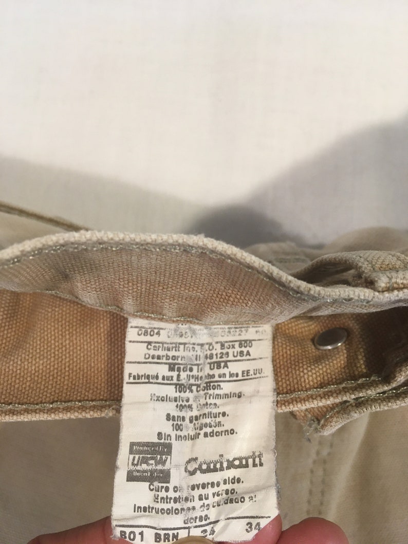 Puede incluir: Una etiqueta de ropa de trabajo Carhartt de color beige con texto blanco. La etiqueta incluye el nombre de la marca, la talla y las instrucciones de cuidado. El texto dice: "0804 08227 Carhartt Inc. P.O. Box 600 Dearborn MI 48126 USA Made in USA Fabriqu&eacute; aux E.U. Hecho en los EE.UU. 100% Cotton. Exclusive Trimming. 100% coton. Sane garniture. 100 algod&oacute;n. Sin incluir adorno.  Procter & Gamble  UREN Carhartt Cure on reverse side. Entretien au verso. Instrucciones de cuidado al dorso. B01 BRN 34"