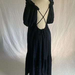 May include: Black maxi dress with a low back and crisscross straps. The dress features short, puffy sleeves and a tiered skirt. The neckline and sleeves are trimmed with a ruffle detail. The dress is displayed on a mannequin against a white backdrop.