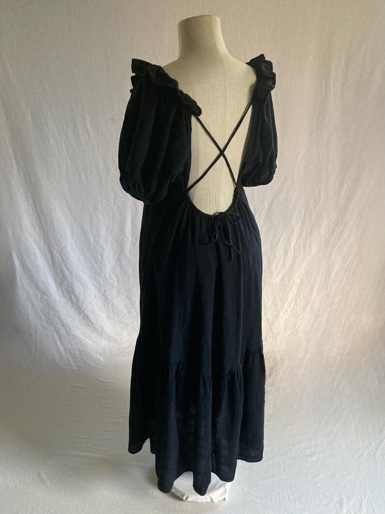 May include: Black maxi dress with a low back and crisscross straps. The dress features short, puffy sleeves and a tiered skirt. The neckline and sleeves are trimmed with a ruffle detail. The dress is displayed on a mannequin against a white backdrop.