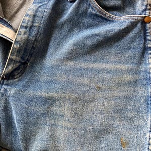 Pu&ograve; includere: Primo piano di jeans di denim blu chiaro con segni di usura. I jeans hanno un classico stile a cinque tasche, rivetti in rame e chiusura con bottone. Il denim mostra sbiadimento e scolorimento, suggerendo un'estetica vintage o usurata.