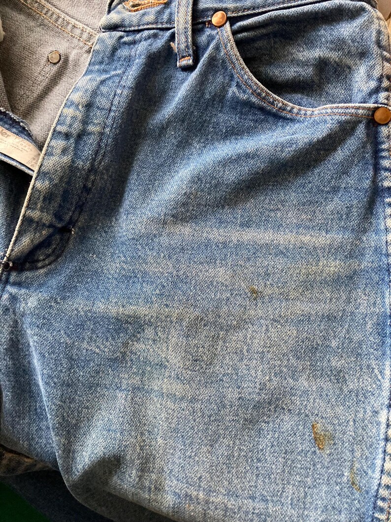 Pu&ograve; includere: Primo piano di jeans di denim blu chiaro con segni di usura. I jeans hanno un classico stile a cinque tasche, rivetti in rame e chiusura con bottone. Il denim mostra sbiadimento e scolorimento, suggerendo un'estetica vintage o usurata.