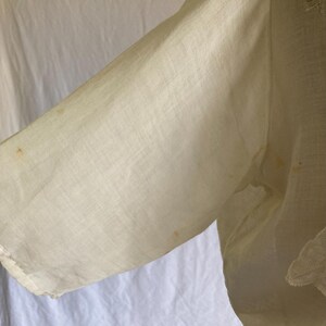 May include: Close-up of a cream-colored garment sleeve. The fabric appears to be a lightweight linen or cotton, with visible texture and slight discoloration. The sleeve is partially visible, with a lace trim detail.