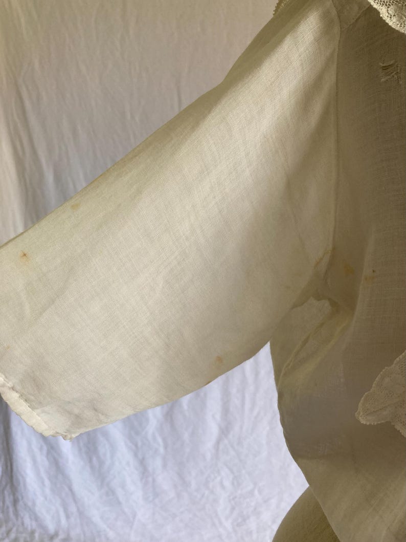 May include: Close-up of a cream-colored garment sleeve. The fabric appears to be a lightweight linen or cotton, with visible texture and slight discoloration. The sleeve is partially visible, with a lace trim detail.