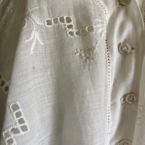May include: Close-up of a white, vintage blouse with intricate embroidery and cutwork details. The blouse features delicate floral and geometric patterns, along with mother-of-pearl buttons. The fabric appears to be linen or a similar lightweight material.