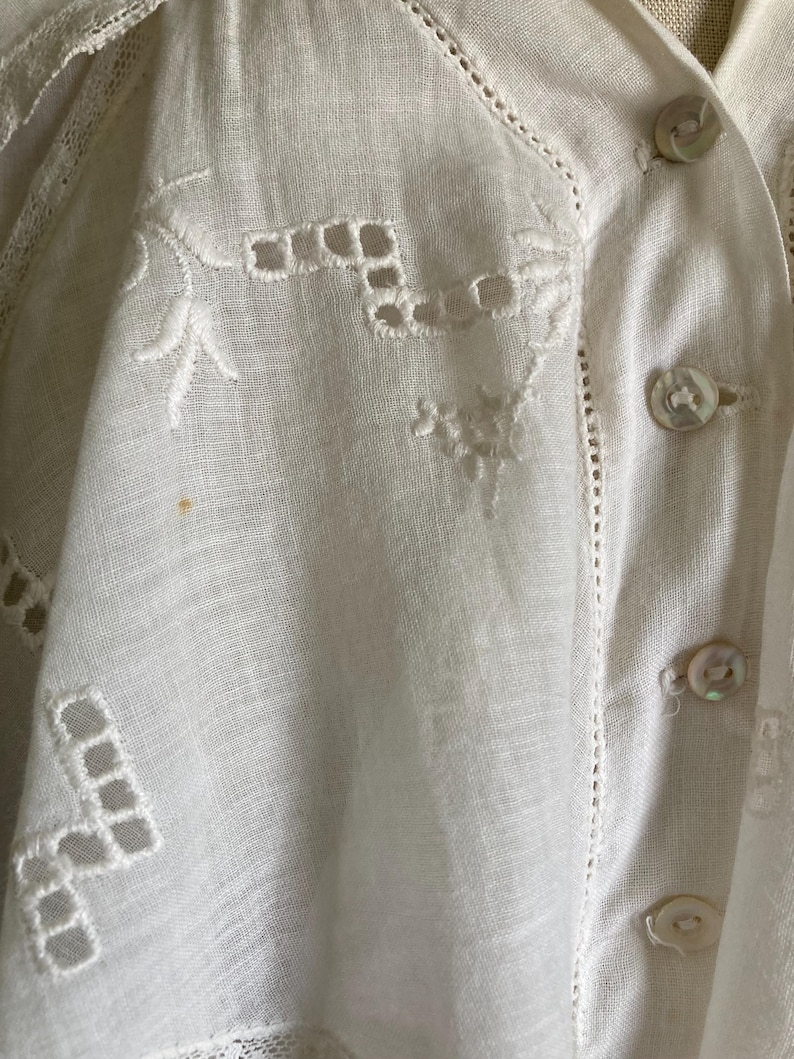 May include: Close-up of a white, vintage blouse with intricate embroidery and cutwork details. The blouse features delicate floral and geometric patterns, along with mother-of-pearl buttons. The fabric appears to be linen or a similar lightweight material.