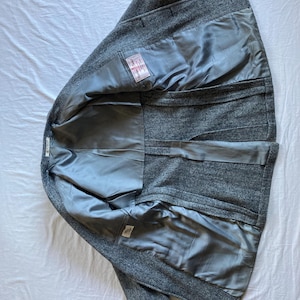 May include: A gray herringbone blazer with a smooth gray lining. The interior features a rectangular white label with red and black text that reads Harris Tweed. The garment is shown from the inside, displaying the construction and fabric.