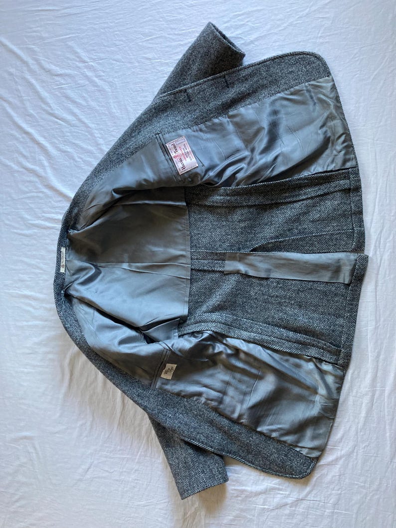 May include: A gray herringbone blazer with a smooth gray lining. The interior features a rectangular white label with red and black text that reads Harris Tweed. The garment is shown from the inside, displaying the construction and fabric.