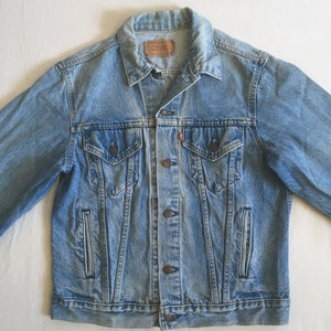 May include: A light blue denim jacket with a button closure, two chest pockets, and two front pockets. The jacket has a faded look and a worn-in feel.
