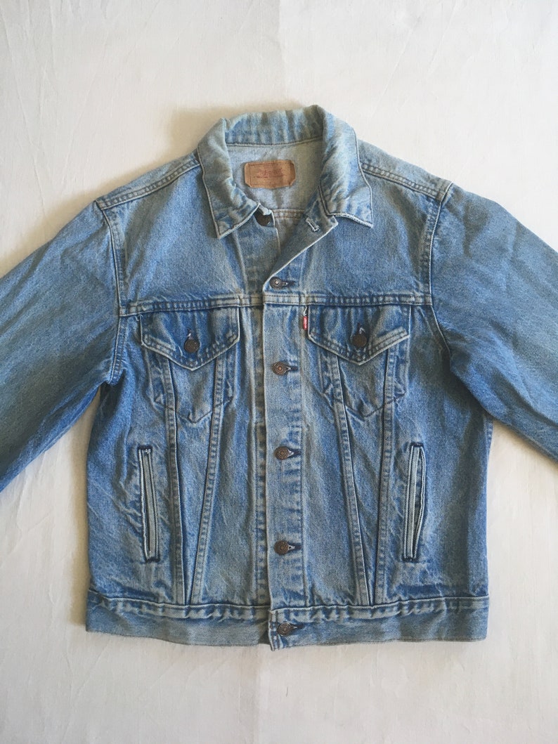 May include: A light blue denim jacket with a button closure, two chest pockets, and two front pockets. The jacket has a faded look and a worn-in feel.