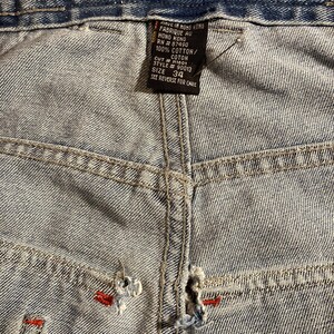K&ouml;nnte beinhalten: Nahaufnahme einer hellblauen Jeans mit Gebrauchsspuren. Die Jeans hat rote N&auml;hte und ein "KIK WEAR"-Etikett in Orange und Rot. Gr&ouml;&szlig;e 34.