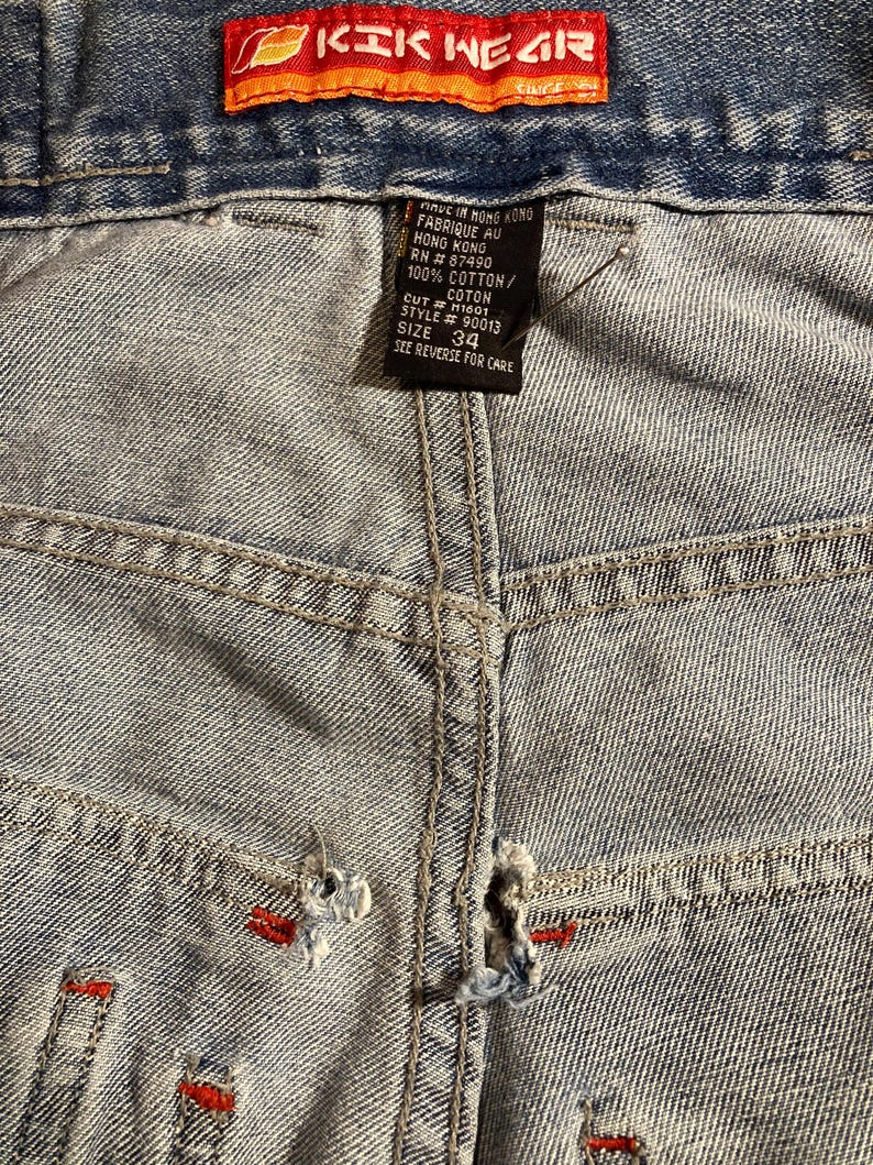 K&ouml;nnte beinhalten: Nahaufnahme einer hellblauen Jeans mit Gebrauchsspuren. Die Jeans hat rote N&auml;hte und ein "KIK WEAR"-Etikett in Orange und Rot. Gr&ouml;&szlig;e 34.