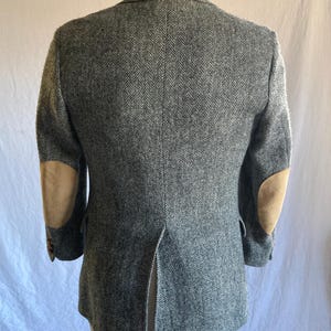 May include: A gray and white herringbone blazer featuring tan suede elbow patches on both sleeves. The jacket has a single center back vent, notched lapels, and long sleeves with buttons at the cuffs, displayed from the back view.