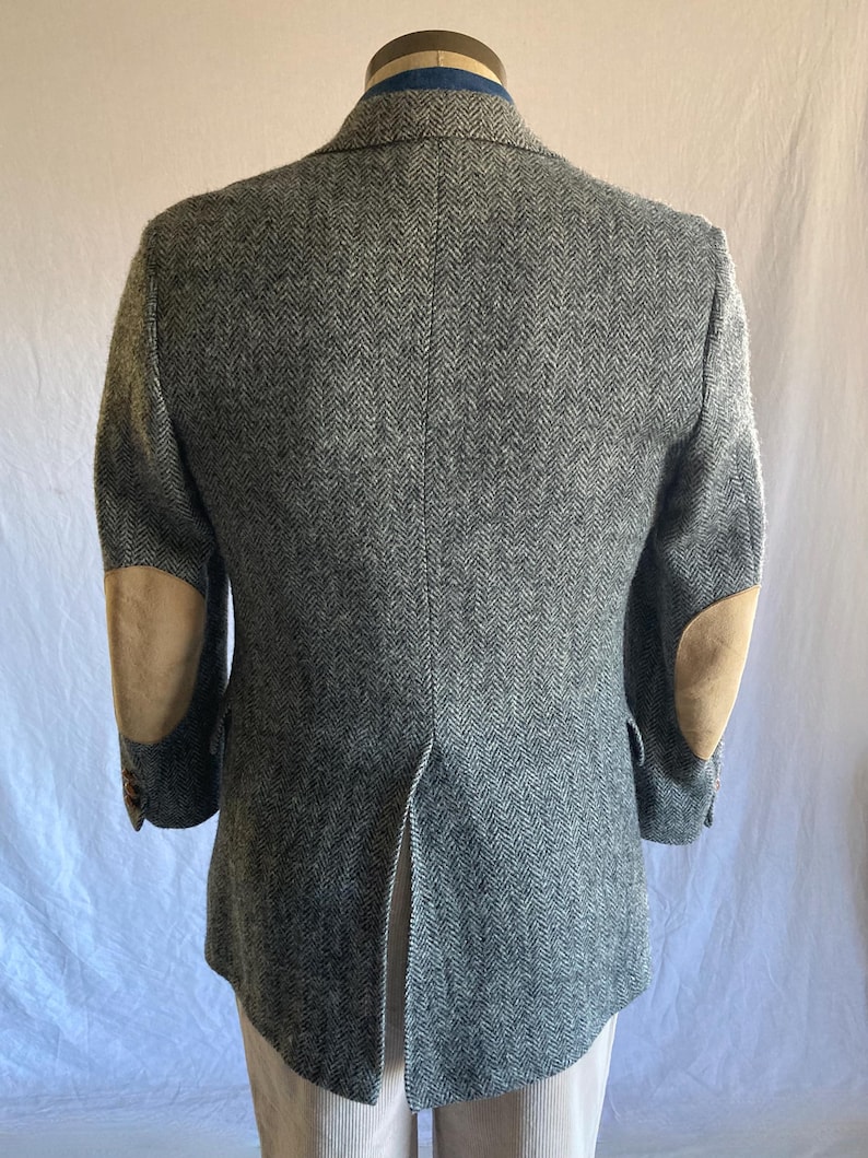 May include: A gray and white herringbone blazer featuring tan suede elbow patches on both sleeves. The jacket has a single center back vent, notched lapels, and long sleeves with buttons at the cuffs, displayed from the back view.
