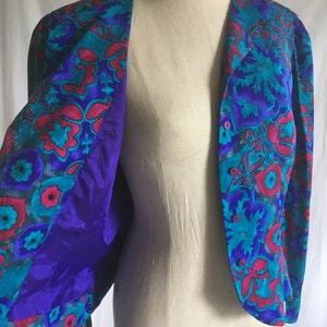 Op de afbeelding: Een paars en turquoise bloemenpatroon jas met een knoopsluiting. De jas is open en de voering is effen paars.
